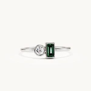 May include: A silver-toned ring featuring a round, clear, brilliant-cut stone set in a bezel and a rectangular, emerald-green, baguette-cut stone set in a vertical orientation. The band is thin, polished, and metallic, connecting both stone settings.