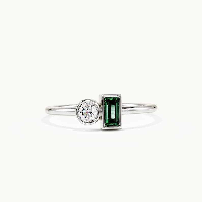 May include: A silver-toned ring featuring a round, clear, brilliant-cut stone set in a bezel and a rectangular, emerald-green, baguette-cut stone set in a vertical orientation. The band is thin, polished, and metallic, connecting both stone settings.