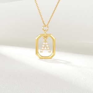 May include: A gold-coloured necklace featuring a rectangular pendant. The pendant has the letter "A" set with small, clear gemstones. The necklace chain is also gold, set against a soft white background.