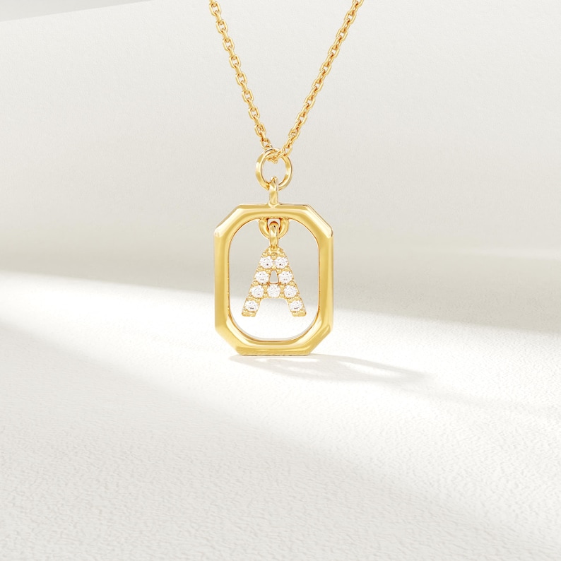 May include: A gold-coloured necklace featuring a rectangular pendant. The pendant has the letter "A" set with small, clear gemstones. The necklace chain is also gold, set against a soft white background.