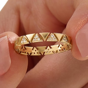 May include: A gold ring with a geometric design featuring alternating triangles. Some triangles are filled with small, sparkling stones. The ring is held between two fingers, showcasing the intricate details and the B14K hallmark.