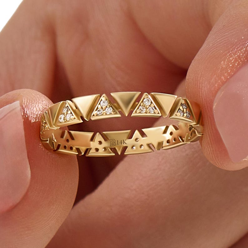 May include: A gold ring with a geometric design featuring alternating triangles. Some triangles are filled with small, sparkling stones. The ring is held between two fingers, showcasing the intricate details and the B14K hallmark.