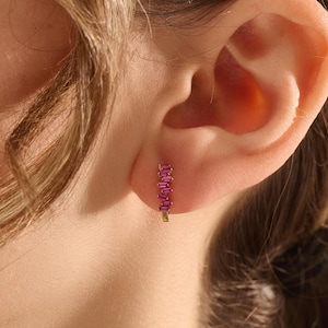 May include: A gold hoop earring with a vertical row of five rectangular, pink gemstones. The earring is worn on an ear, with the focus on the jewelry. The gemstones are a vibrant pink color.