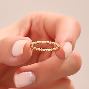 May include: A gold ring featuring a delicate beaded design. The ring is held between fingers with manicured nails. The ring's design consists of small, evenly spaced beads around the entire band. The ring is a simple yet elegant piece of jewelry.