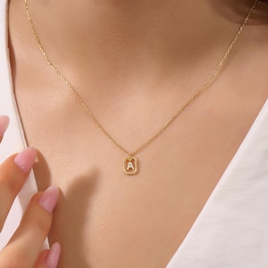 May include: A gold necklace with a small rectangular pendant. The pendant has the letter "A" in white. The necklace is worn around the neck.