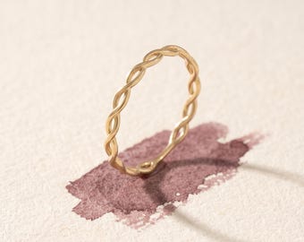 May include: A thin, gold-toned ring featuring a continuous twisted rope or braided metal design. The band has a smooth, polished finish and a circular shape, standing upright to showcase the intricate, repeating pattern of the metal wire.
