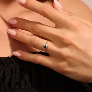 May include: A gold ring featuring a square, dark blue gemstone, accented by smaller white and light blue stones. The ring is set with a delicate band and is shown on a person's finger.