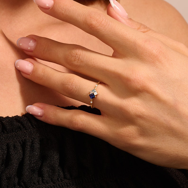 May include: A gold ring featuring a square, dark blue gemstone, accented by smaller white and light blue stones. The ring is set with a delicate band and is shown on a person's finger.