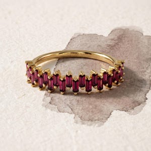 May include: A gold ring featuring a series of vertical, baguette-cut deep red gemstones set along the band. The interior of the gold band is clearly stamped with the text 14K, indicating the metal purity of this jewelry piece.