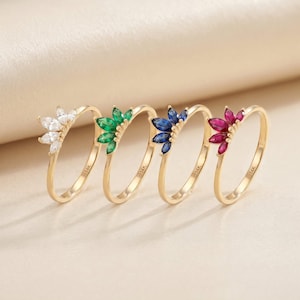 Könnte beinhalten: Vier goldene Ringe mit Edelstein-Clustern in Weiß, Grün, Blau und Rot. Jeder Ring hat ein dünnes Band und eine Ansammlung von Edelsteinen, die in einem floralen Muster angeordnet sind. Die Ringe werden auf einer hellbeige Oberfläche präsentiert.