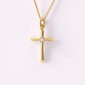May include: A gold-colored cross pendant necklace. The cross features a small, clear gemstone at the center. The necklace chain is also gold-colored and hangs against a white background. The cross is a symbol of faith and spirituality.