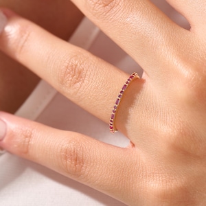 May include: A gold-colored ring with a band of small, rectangular, red gemstones. The ring is worn on a finger, with the hand and part of the arm visible in the image. The ring's design is simple and elegant.