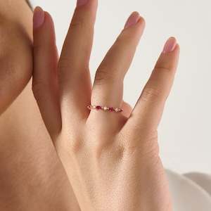May include: A thin gold band ring featuring an alternating pattern of marquise-cut deep pink gemstones and small round clear stones. The delicate metal band holds the stones in a horizontal row, creating a continuous, refined, and elegant design.