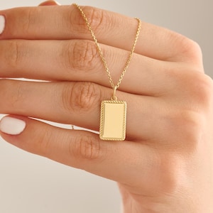 May include: A gold-toned necklace featuring a rectangular pendant with a smooth, flat centre and a textured, rope-style border. The pendant hangs from a delicate, matching gold-toned chain, creating a simple and refined piece of jewellery.