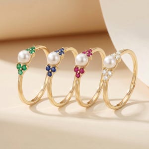 May include: Four gold rings with a pearl and gemstone cluster design. Each ring features a gold band and a cluster of green, blue, red, or white gemstones. The pearls are white and the gemstones are emerald, sapphire, ruby, and diamond.