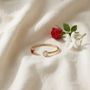 May include: A gold open cuff bracelet with a red gemstone and a clear, round gemstone. The bracelet is on a white, textured surface, with a red rose and white jasmine flowers. The bracelet is a stylish piece of jewellery.