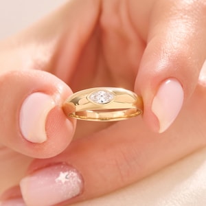 May include: A polished gold band ring featuring a single marquise-cut clear gemstone set horizontally in the centre. The metal surface is smooth and rounded, creating a sleek profile that highlights the faceted stone embedded within the band.