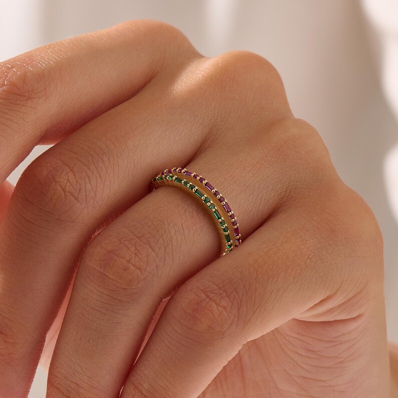 Op de afbeelding: Een gouden ring met twee rijen edelstenen. E&eacute;n rij heeft kleine, rechthoekige groene stenen en de andere rij heeft kleine, rechthoekige rode stenen. De ring zit om de vinger van een persoon.