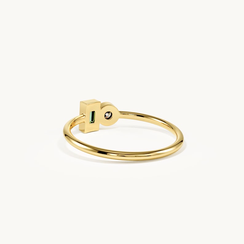May include: A gold ring featuring a thin, polished band. The focal point consists of a rectangular setting containing a small, green, baguette-cut gemstone positioned next to a circular setting holding a single, clear, round-cut gemstone.