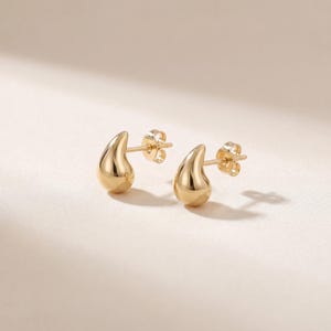May include: A pair of gold-colored teardrop stud earrings. The earrings have a smooth, polished finish and a post-and-butterfly-clasp closure. The earrings are displayed on a light-colored surface.