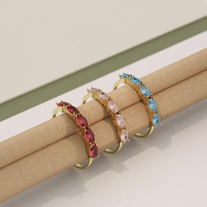 Pode incluir: Três anéis de ouro, cada um com uma fileira de pedras preciosas ovais. Os anéis apresentam pedras vermelhas, rosa e azuis. Os anéis são exibidos em uma superfície marrom clara contra um fundo branco e verde.