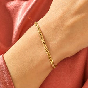 May include: A gold-toned metal chain bracelet featuring a classic curb link design. The bracelet is worn around the wrist, showcasing a polished, reflective surface and a uniform, interlocking pattern of small, rounded links in a bright yellow gold hue.