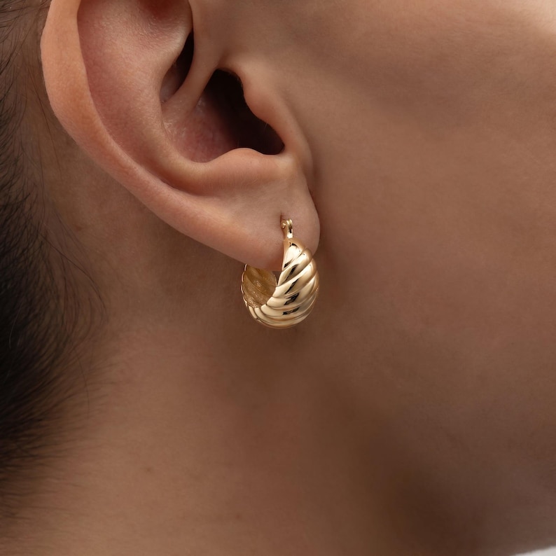 May include: A close-up of a gold hoop earring with a textured, ridged design. The earring is attached to an ear. The earring has a shiny, metallic finish and a classic, timeless style.