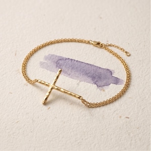 Op de afbeelding: Een goudkleurige armband met een dubbel kettingontwerp en een centrale kruishanger met een gehamerd, getextureerd oppervlak. Het kruis is geïntegreerd in de ketting, die is voorzien van een kleine karabijnsluiting en een verstelbaar verlengstuk.