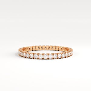 May include: A rose gold eternity ring with a continuous row of round-cut, clear gemstones. The ring is set against a white background, highlighting the sparkle of the stones. This elegant piece is a classic jewelry item.
