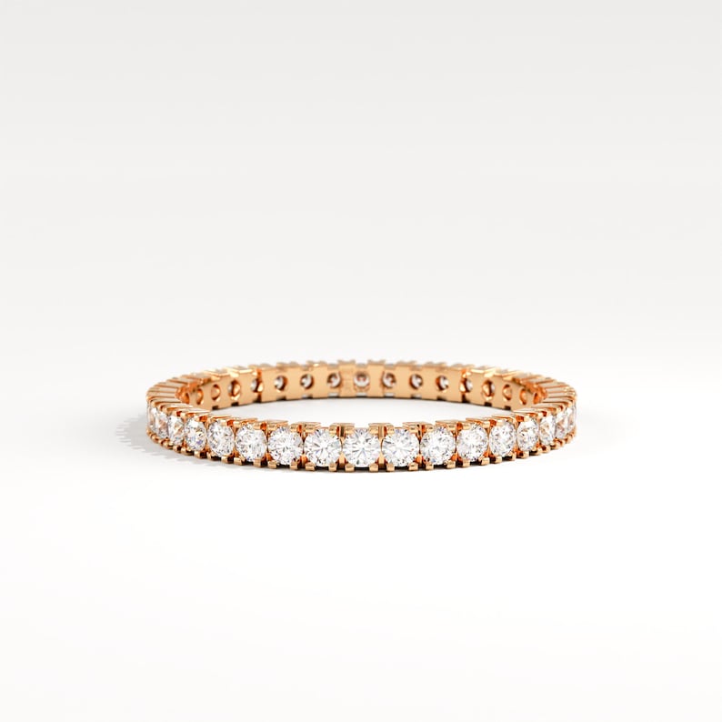 May include: A rose gold eternity ring with a continuous row of round-cut, clear gemstones. The ring is set against a white background, highlighting the sparkle of the stones. This elegant piece is a classic jewelry item.