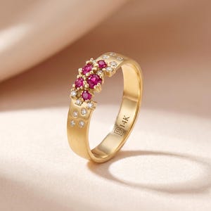 May include: A gold ring with a cluster of small, round, red gemstones and small, clear gemstones. The ring has a smooth, matte finish and is marked with "B14K". The gemstones are set along the top of the ring.