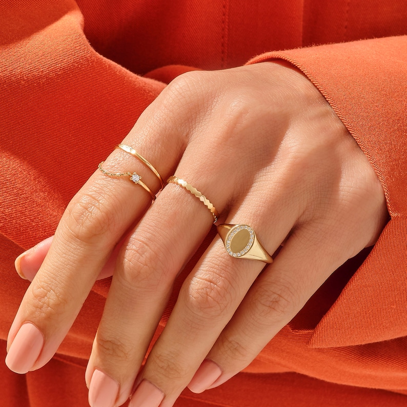 May include: A hand wearing four gold rings. The ring on the index finger is a signet ring with a clear oval stone surrounded by smaller stones. The other rings are thin and delicate.