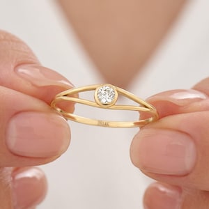 May include: A gold ring with a single, round, clear gemstone set in the center. The ring has a split band design. The inscription "B14K" is visible on the band. The ring is held between two hands.