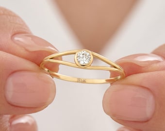 May include: A gold ring with a single, round, clear gemstone set in the center. The ring has a split band design. The inscription "B14K" is visible on the band. The ring is held between two hands.