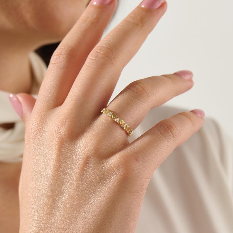 May include: A gold ring with a geometric design featuring small, raised pyramid shapes and interspersed with small, clear gemstones. The ring is worn on a finger, showcasing its intricate details and elegant style.