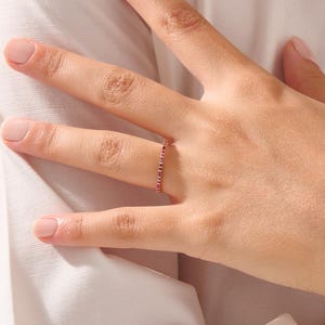 Op de afbeelding: Een delicate ring met kleine, afwisselende rode en donkerblauwe edelstenen. De ring wordt om een vinger gedragen en toont de fijne details en het elegante ontwerp. De edelstenen zijn dicht op elkaar geplaatst, waardoor een doorlopende lijn van kleur ontstaat.