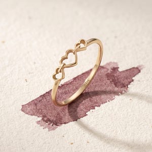 May include: A gold ring featuring three connected open heart shapes along the top band. The interior of the band is clearly stamped with the text 14K, indicating the metal purity of this delicate piece of jewellery.