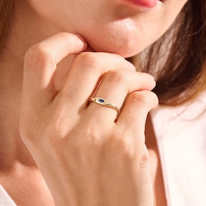 May include: A gold band ring featuring a single marquise-cut blue gemstone set horizontally in the center. The ring has a smooth, polished finish and a tapered design that narrows toward the back of the band, showcasing a simple and clean aesthetic.