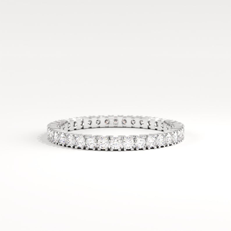 May include: A silver eternity ring with a continuous row of round-cut, clear gemstones. The ring is set against a white background, showcasing the sparkle of the stones. A classic piece of jewelry.