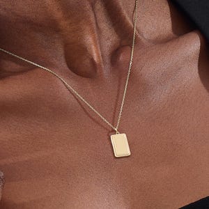 May include: A gold-toned necklace featuring a delicate chain and a rectangular pendant. The pendant has a smooth, flat centre surface framed by a textured, rope-like border. The necklace rests against the skin, showcasing the metallic finish.