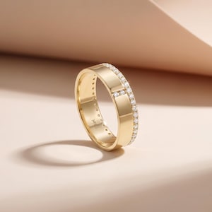 May include: A gold ring with a band of small, clear gemstones set along the top and around the edges. The ring is made of polished gold and has a smooth, flat surface. The ring is set against a soft, neutral background.