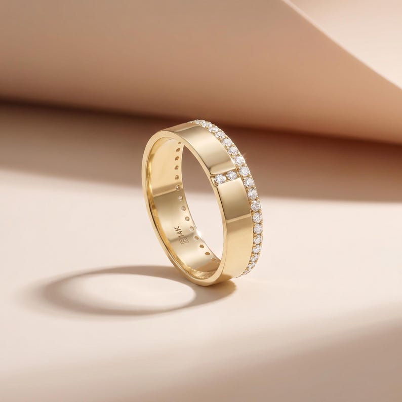 May include: A gold ring with a band of small, clear gemstones set along the top and around the edges. The ring is made of polished gold and has a smooth, flat surface. The ring is set against a soft, neutral background.