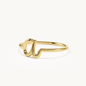 May include: A polished gold ring featuring a continuous, smooth metal band that forms the lowercase letter 'a' as the central focal point. The design is crafted from a single piece of rounded gold wire, creating a fluid and seamless aesthetic.