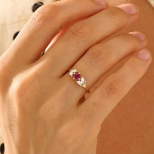 Könnte beinhalten: Ein Goldring mit einem zentralen quadratischen roten Rubin, der auf jeder Seite von zwei kleinen weißen Diamanten im Marquise-Schliff flankiert wird. Die Ringschiene besteht aus poliertem Gelbgold und zeigt ein filigranes Design.