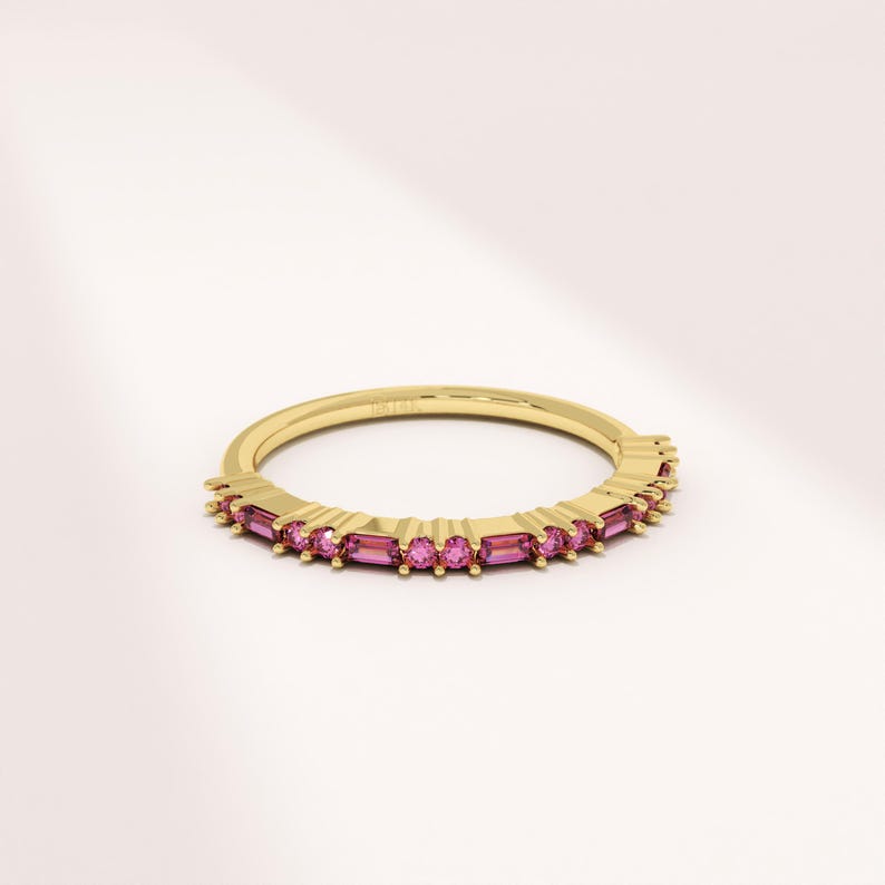 Op de afbeelding: Een gouden ring met een band van afwisselende roze rechthoekige en ronde edelstenen. De ring is geplaatst tegen een witte achtergrond, met zachte verlichting.