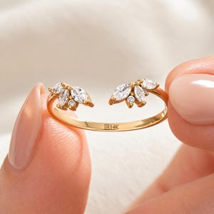 Op de afbeelding: Een open gouden ring met aan beide uiteinden twee clusters van marquise- en rondgeslepen heldere stenen. De binnenkant van de ring is duidelijk gegraveerd met de tekst 14K, wat de zuiverheid van het metaal aangeeft.