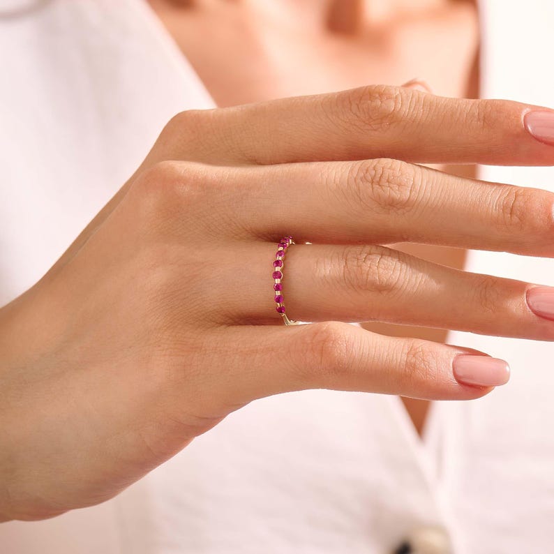May include: A thin gold band ring featuring a series of small, round, vibrant pink gemstones set in a row along the top half of the band. The ring is worn on a finger, showcasing the delicate metalwork and the bright, saturated colour of the stones.