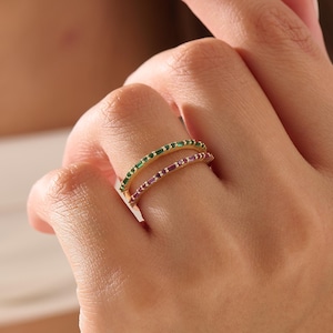 Op de afbeelding: Twee dunne gouden ringen die samen om een vinger worden gedragen. De bovenste ring is voorzien van een rij kleine, rechthoekige groene edelstenen, terwijl de onderste ring is bezet met een rij kleine, rechthoekige roze edelstenen.