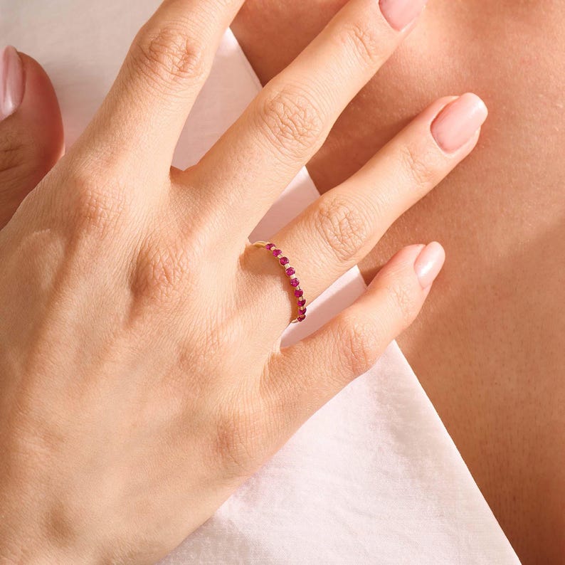 May include: A thin gold band ring featuring a series of small, round, vibrant pink gemstones set along the top half of the band. The ring is worn on a finger, showcasing the delicate metalwork and the consistent size and colour of the stones.