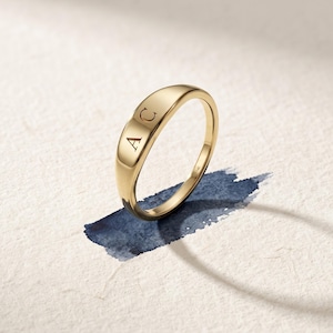 May include: A polished gold signet ring featuring a smooth, rounded top surface engraved with the capital letters A and C. The interior of the band is clearly marked with the stamp 14K, indicating the metal purity of this yellow gold jewelry piece.